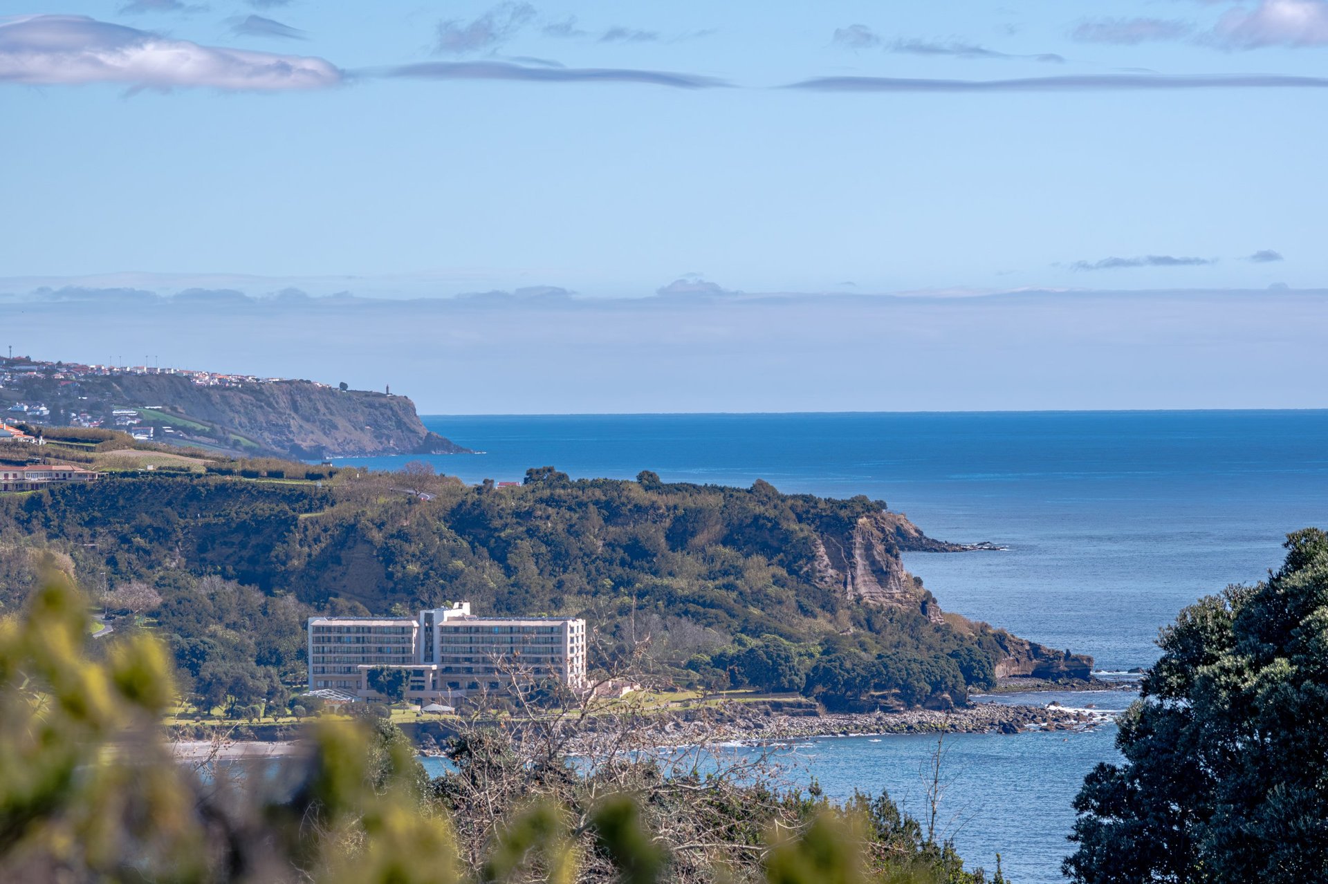 View from the Miradouro do Pisão looking east toward Praia de Água D'Alto, Grande, and the Pestana Bahia Praia Hotel. Framed by trees in the foreground, the blue sea and sky in the background.