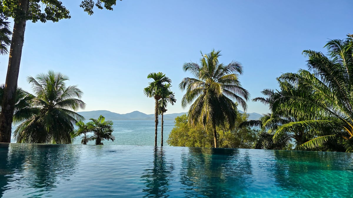 A stunning infinity pool blends seamlessly with the turquoise waters, surrounded by tropical palm trees and mountains