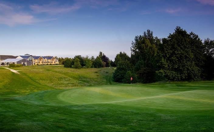 Golf course with manicured greens, tree-lined fairway, and residential clubhouse in background under blue sky