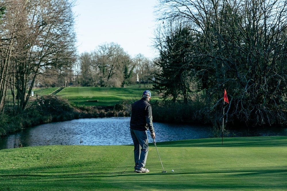 Golfer putting on green near water hazard with trees and red flag in background