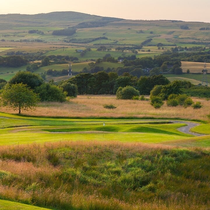 Rolling green hills with fields, trees, and a winding stream in a scenic rural valley landscape
