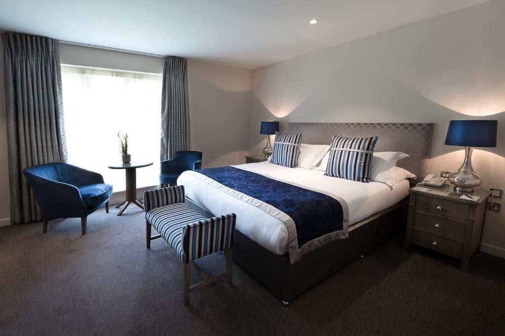 Modern hotel bedroom with king bed, blue accent chair, striped cushions, nightstands with lamps, and large window with curtains