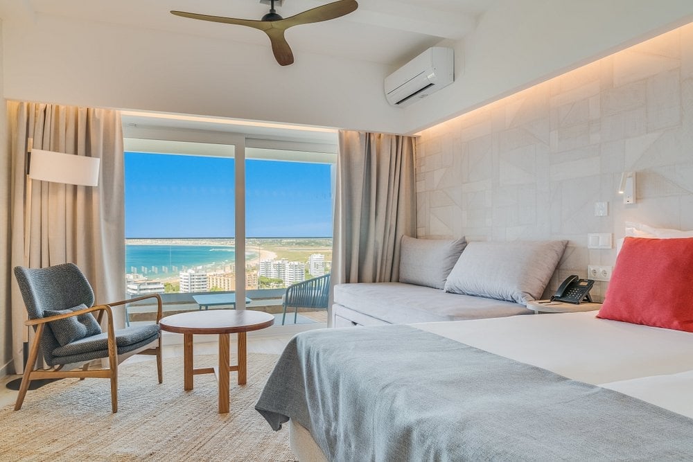 Modern hotel bedroom with twin beds, ocean view from floor-to-ceiling windows, beige decor, and comfortable seating area