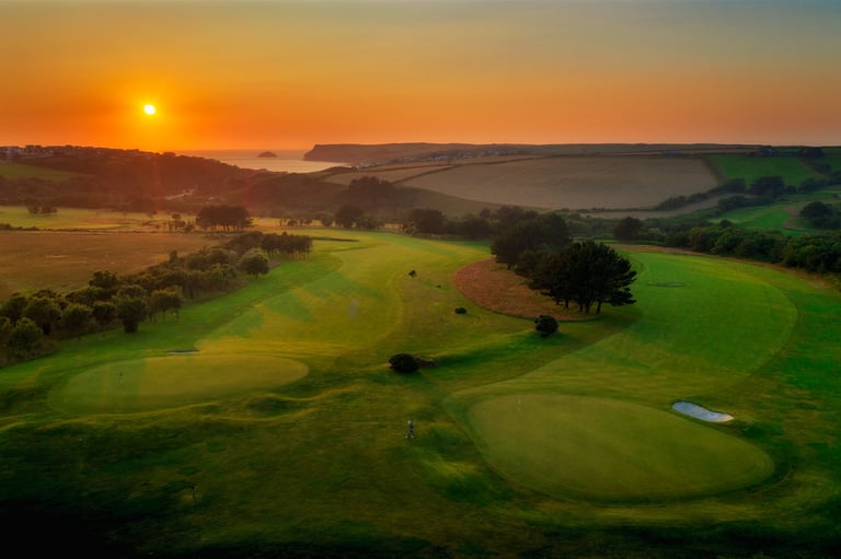 Aerial view of a golf course at sunset with vibrant green fairways, bunkers, and trees under a golden sky