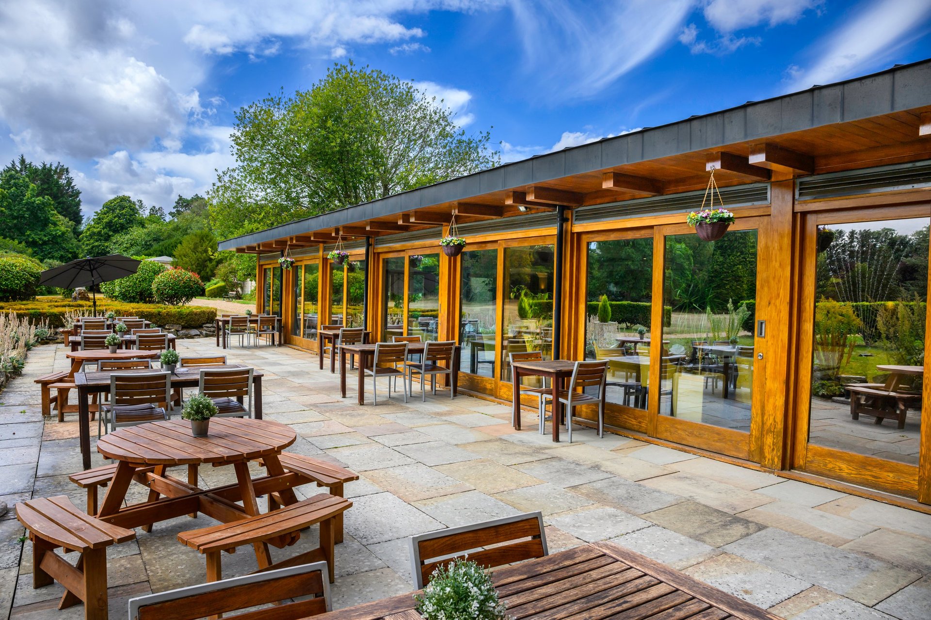 Modern outdoor pavilion with wooden picnic tables on a stone patio, surrounded by green landscaping and blue sky