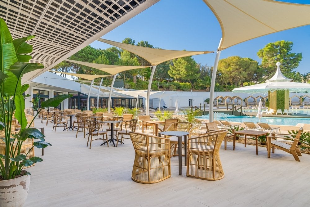 Modern poolside patio with woven dining chairs, cream-colored shade sails, lush greenery, and sparkling swimming pool in bright daylight.