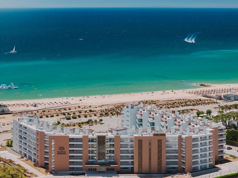 Aerial view of beachfront resort complex with multi-story apartment buildings, sandy beach with umbrellas, and turquoise Mediterranean waters with sailing boats