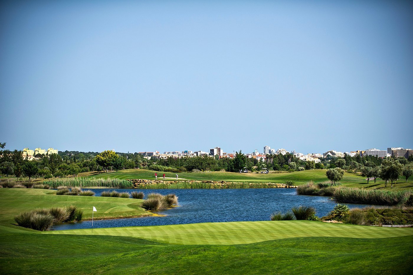 Scenic golf course with blue water hazard, manicured green grass, and city skyline in the background