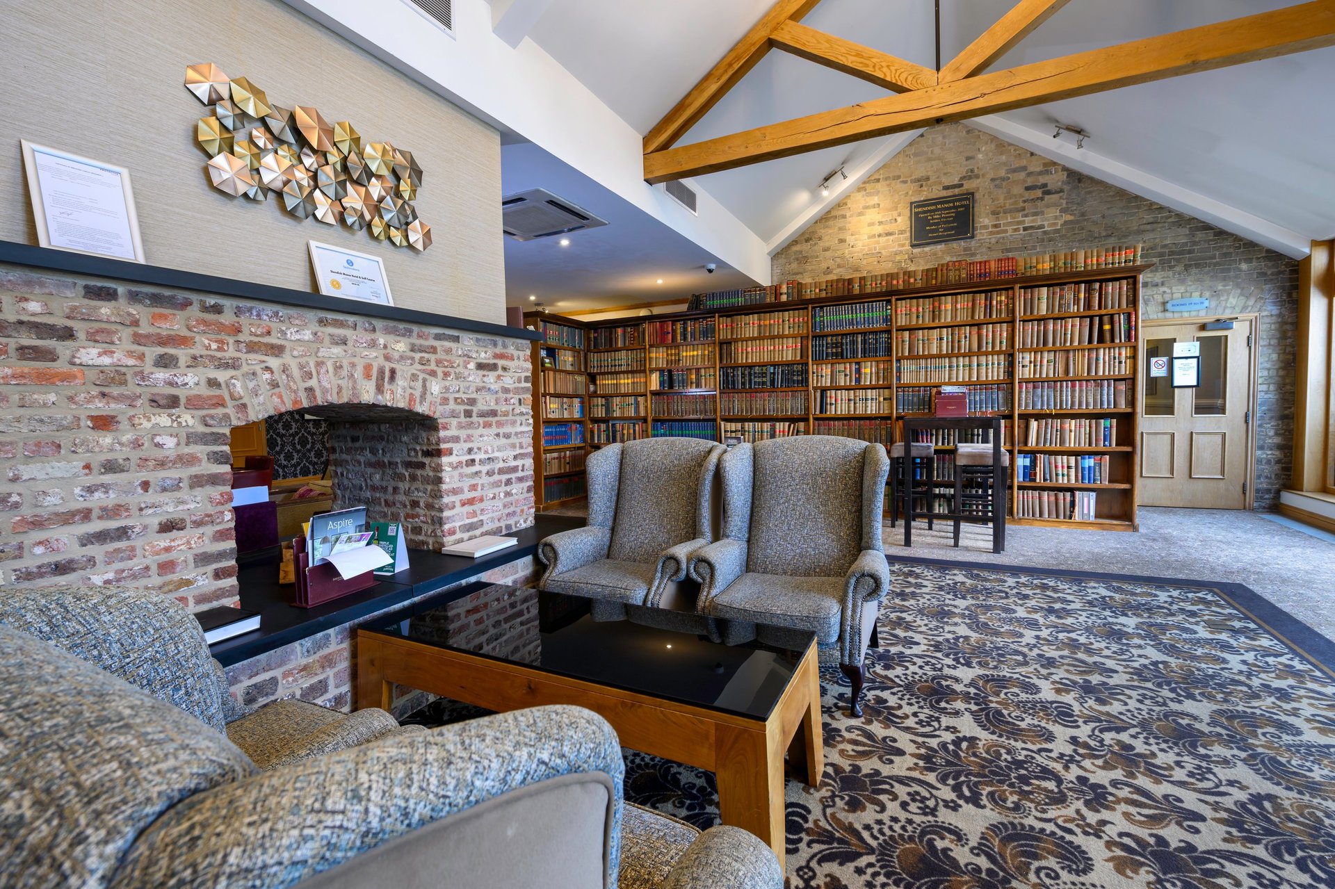 Modern living room with fireplace, bookshelves, upholstered seating, wooden beams, and exposed brick walls