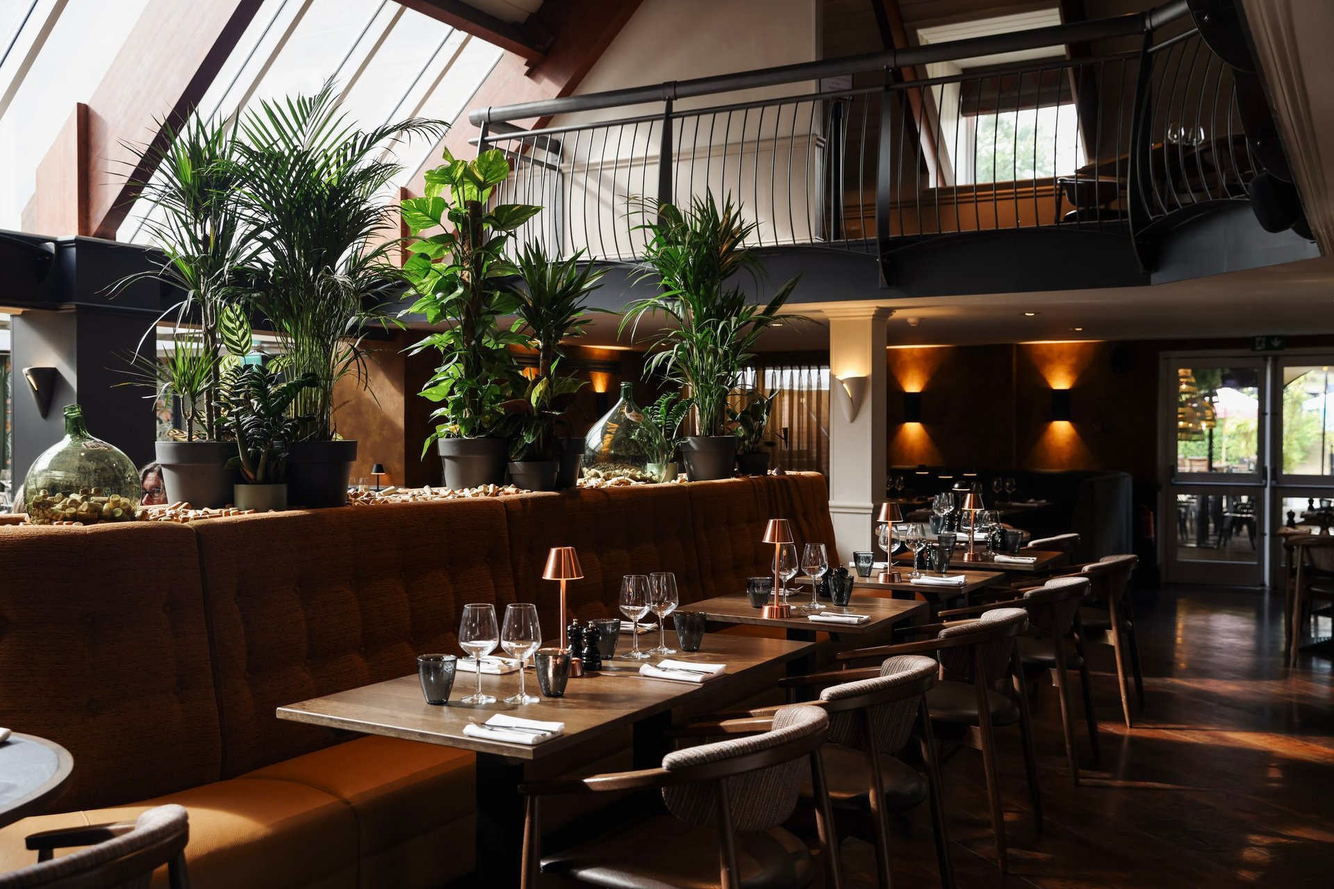 Modern restaurant interior with long wooden dining table, black chairs, potted plants, and skylight overhead