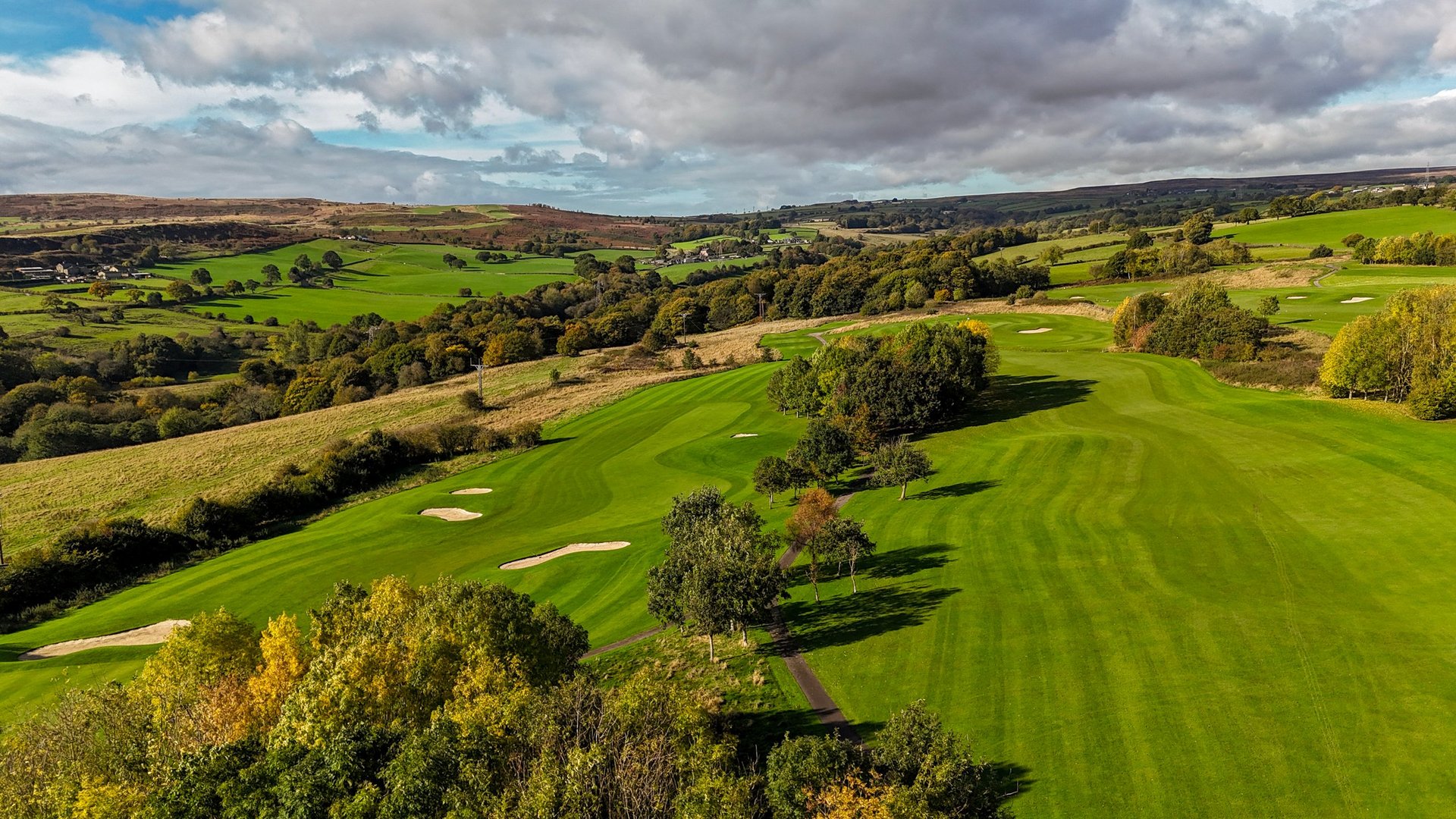 Aerial view of a lush green golf course with manicured fairways, scattered trees, and rolling countryside under cloudy sky