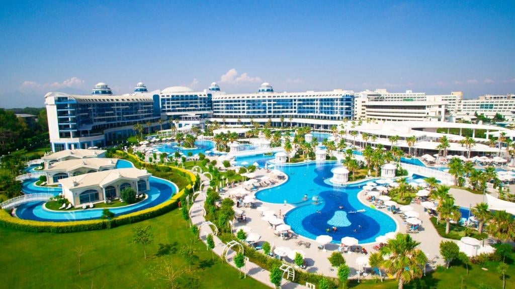 Large resort complex with multiple blue swimming pools, surrounding buildings, manicured lawns, and tropical landscaping