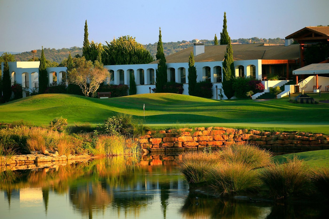 Modern Mediterranean villa with blue walls and cypress trees beside a reflective pond with manicured green lawn