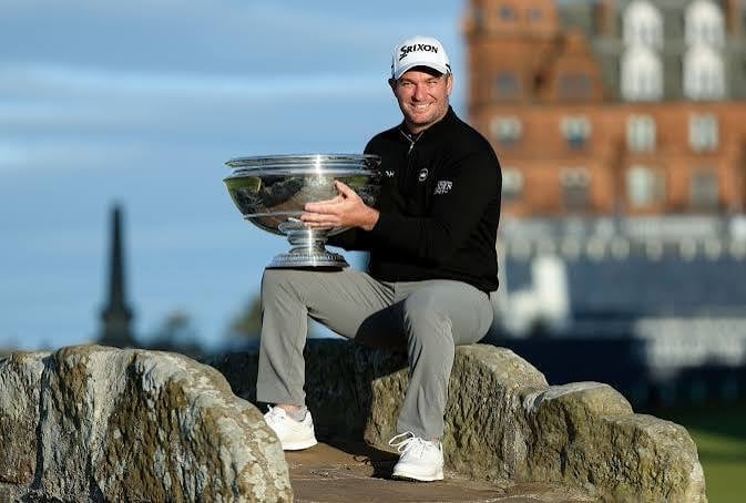 Golfer in white cap and black jacket holds championship trophy while sitting on rocks, with historic building in background