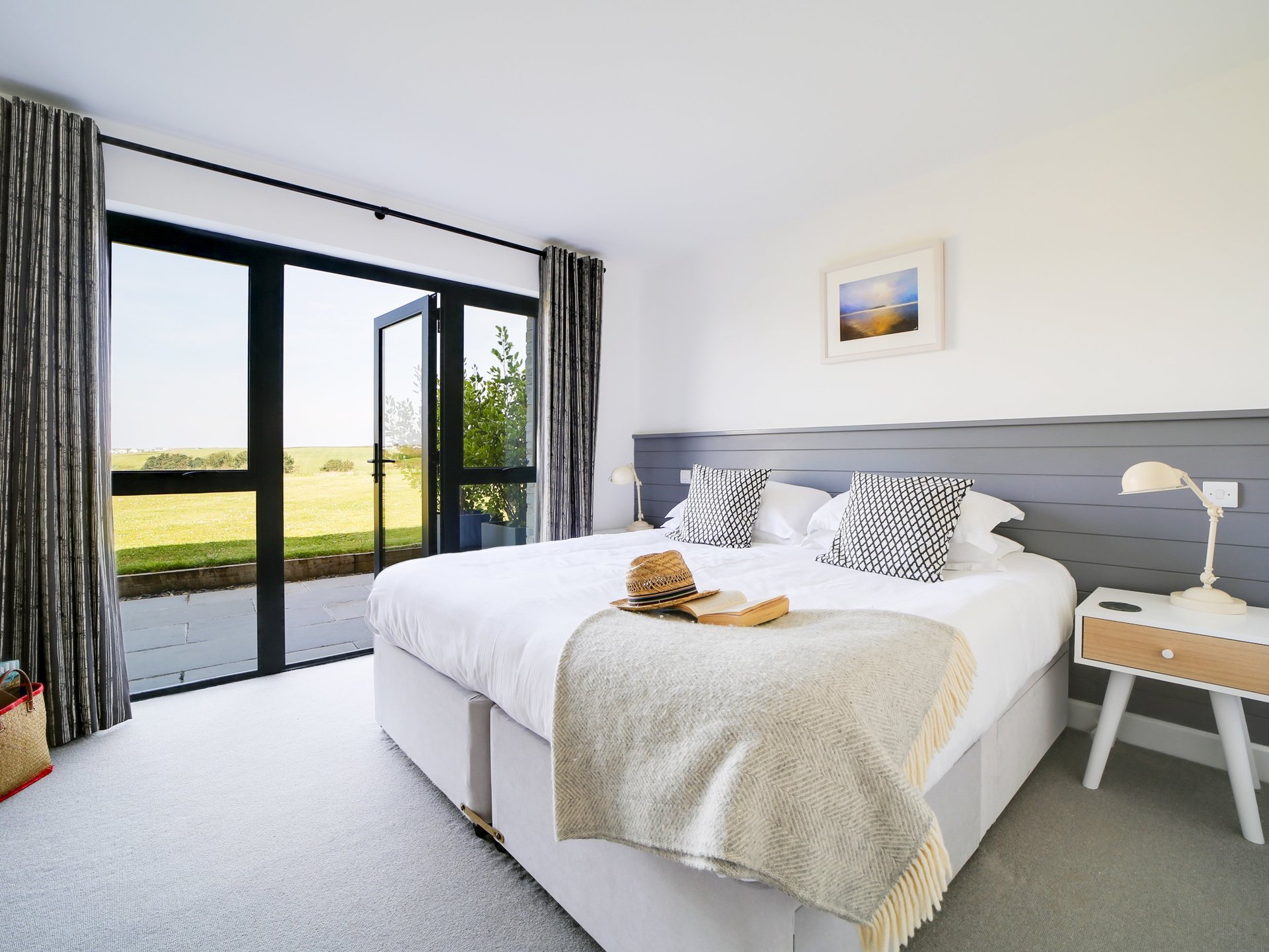 Modern bedroom with white bedding, gray headboard, glass patio doors overlooking green landscape, and minimalist furnishings