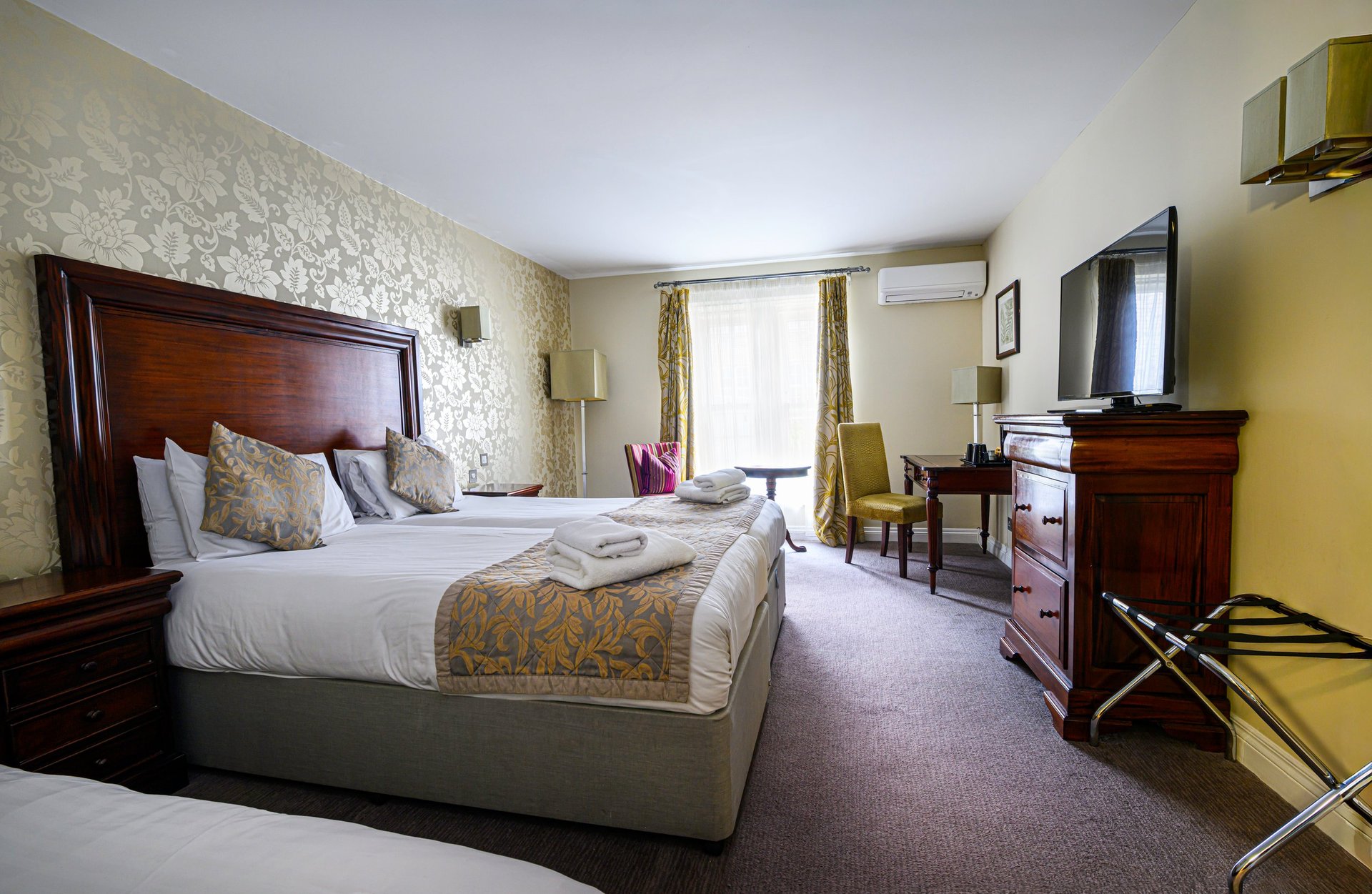Spacious hotel bedroom with wooden bed frame, patterned wallpaper, sitting area, and flat-screen TV