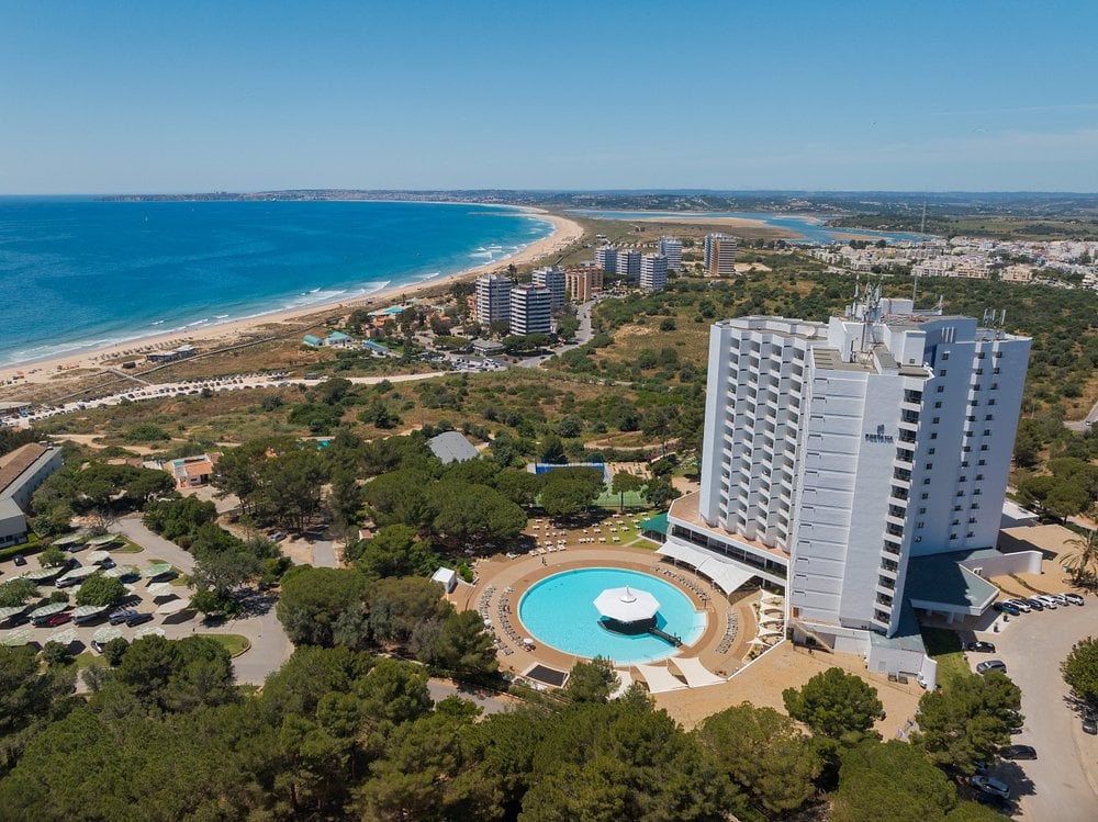 Aerial view of a beachfront resort with a high-rise hotel, circular pool, sandy beach, and Mediterranean coastline