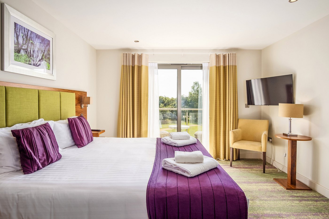 Modern hotel bedroom with king bed, lime green headboard, purple pillows and bench, gold curtains, flat-screen TV, and seating area with view of outdoor landscape