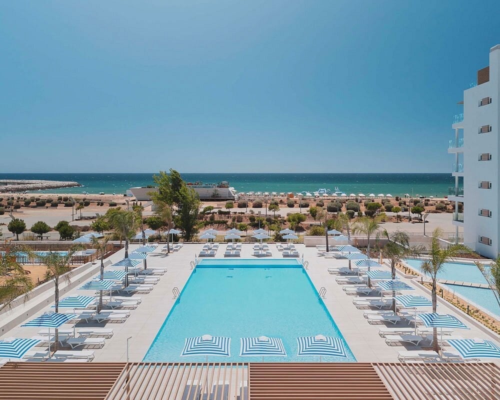 Aerial view of a resort pool overlooking a beach with striped loungers, palm trees, and turquoise Mediterranean Sea