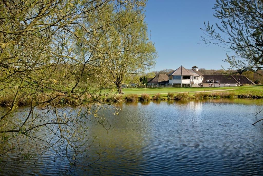 Rural farmhouse with barn beside calm river, framed by green willow trees under blue sky