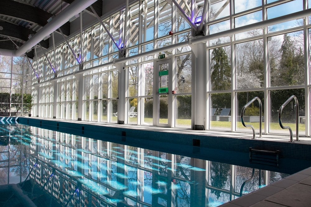 Modern indoor swimming pool facility with large windows, blue tiled walls, and natural light flooding through expansive glass panels overlooking outdoor scenery.