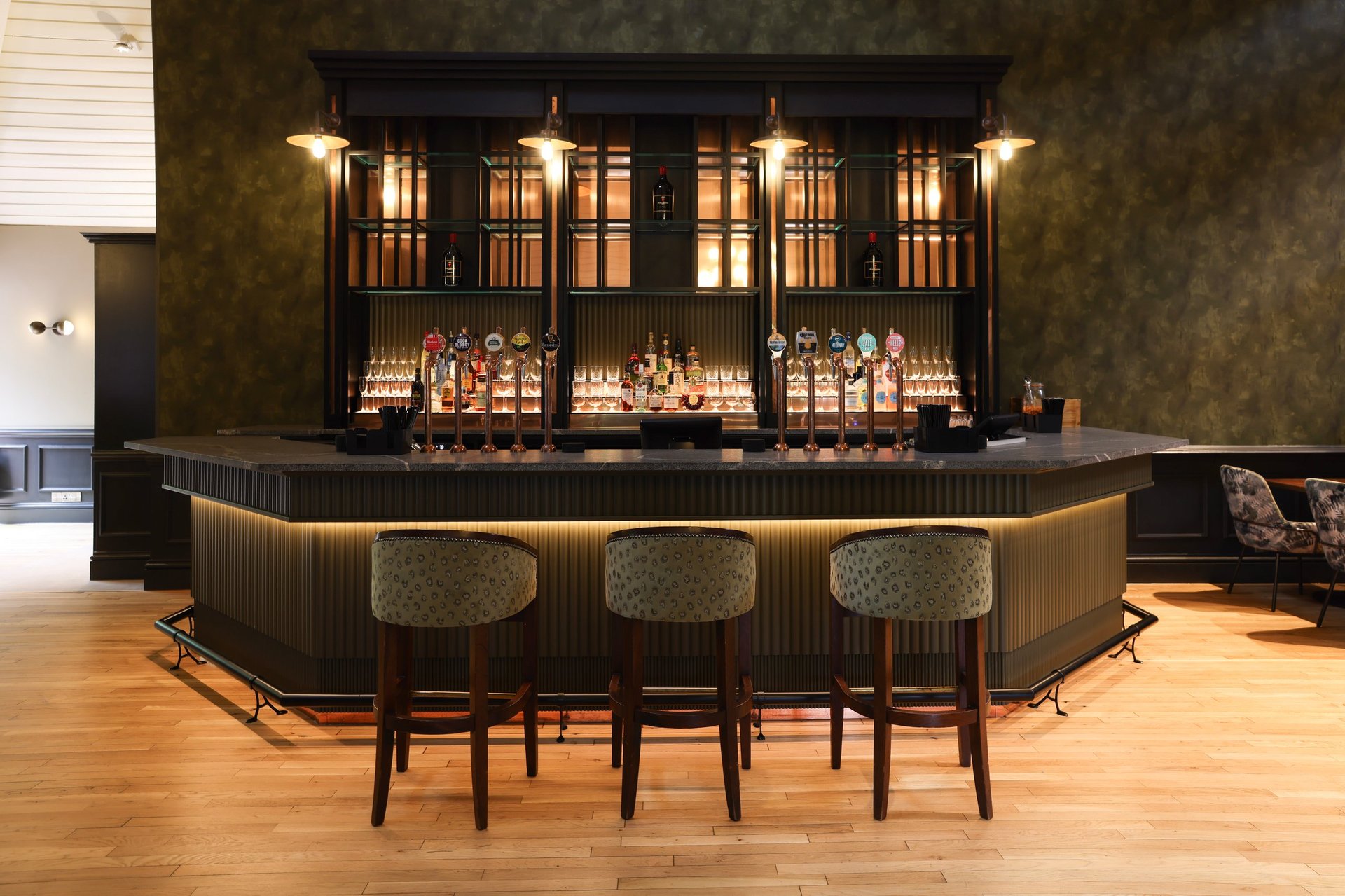Modern upscale bar with curved dark wood counter, three cushioned stools, illuminated display shelves with bottles, and warm lighting