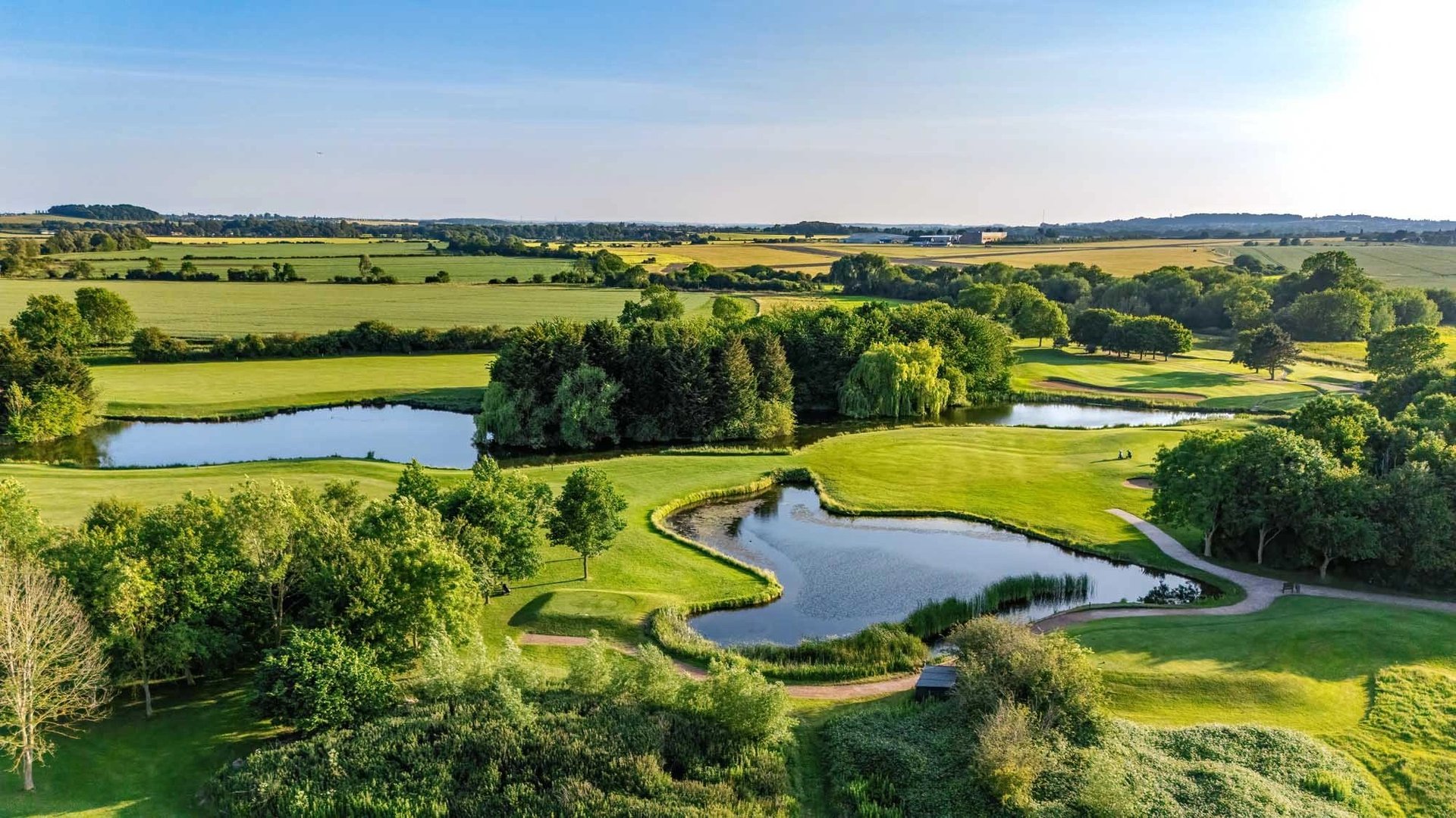Aerial view of green rolling countryside with winding ponds, trees, and farmland under blue sky