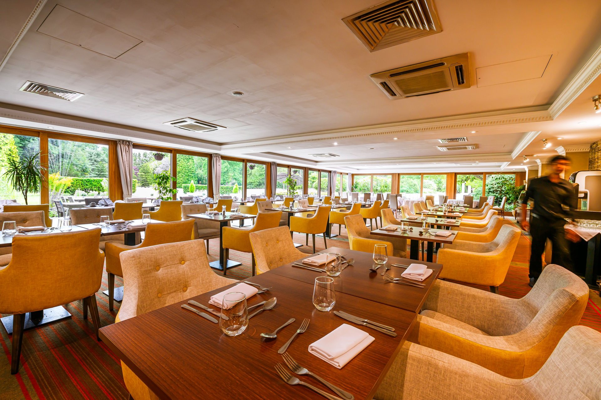 Modern restaurant or hotel dining room with golden upholstered chairs, wooden tables, large windows overlooking greenery, and contemporary ceiling fixtures.
