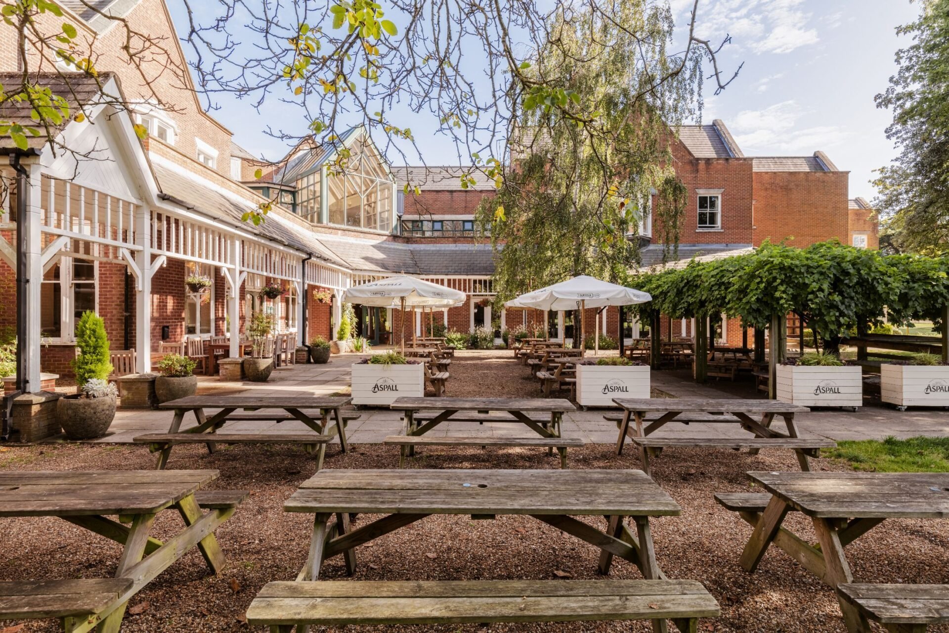 Outdoor beer garden with wooden picnic tables, umbrellas, and historic brick buildings surrounding a paved courtyard