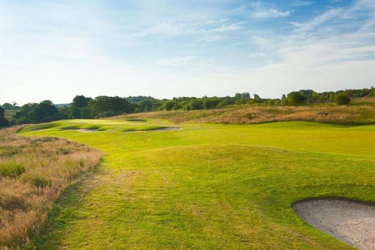 Golf course fairway with green grass, sand bunker, scattered trees, and blue sky with clouds