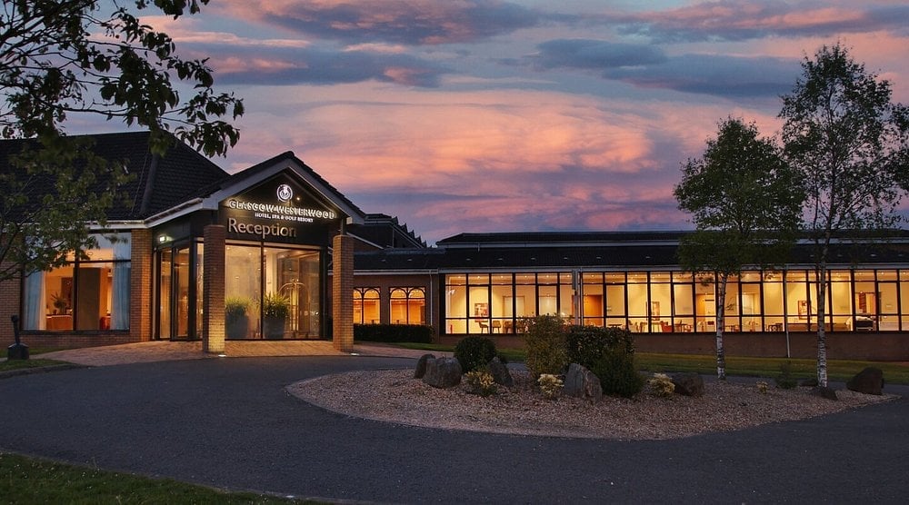 Modern building with illuminated windows at dusk, surrounded by landscaping and trees against a colorful twilight sky