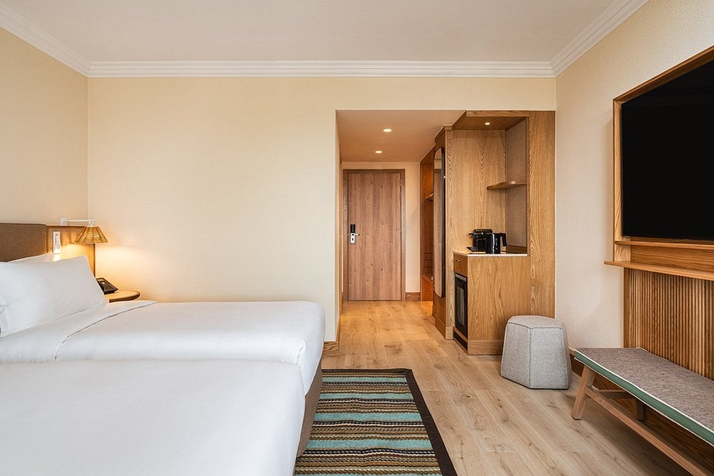 Modern hotel room with white bed, wood furnishings, flat-screen TV, and open bathroom visible in background