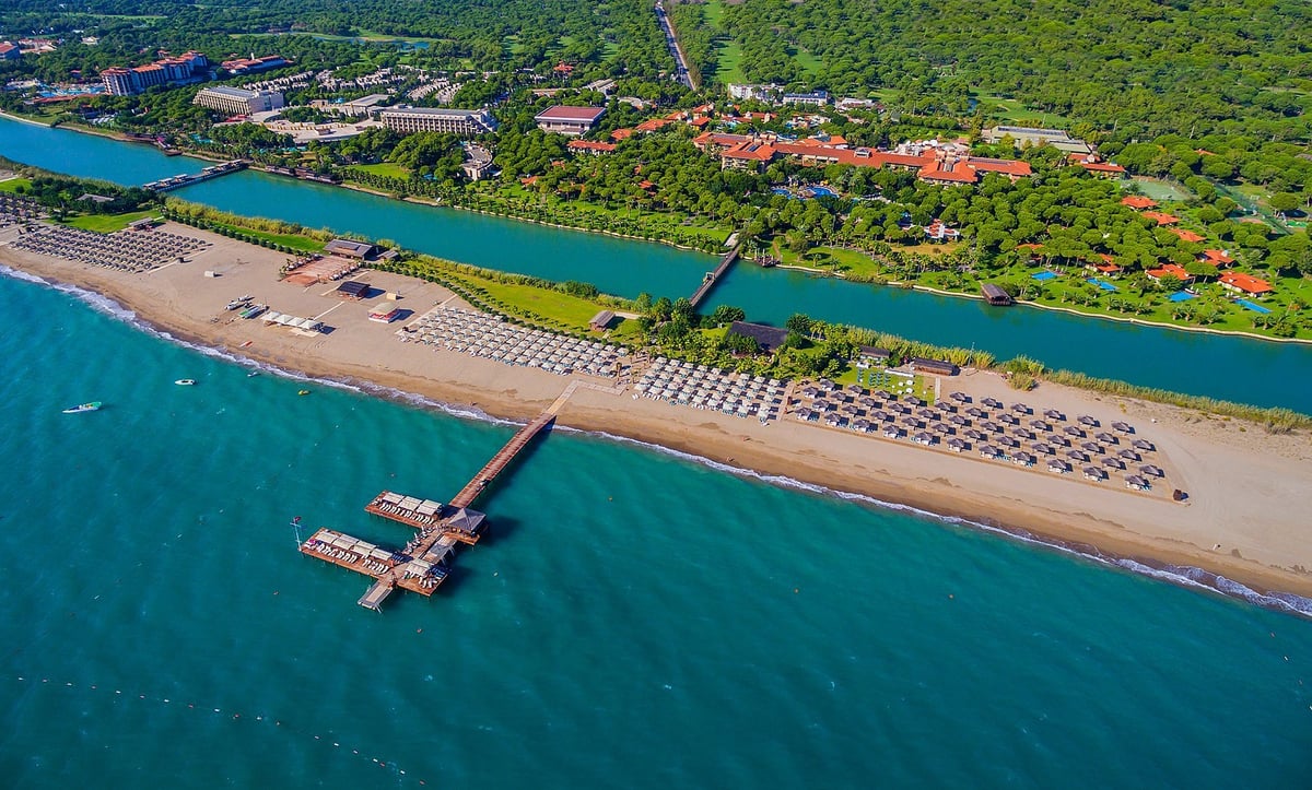 Aerial view of a coastal beach with a pier extending into turquoise water, lined with beach umbrellas, bordered by lush green vegetation and a river