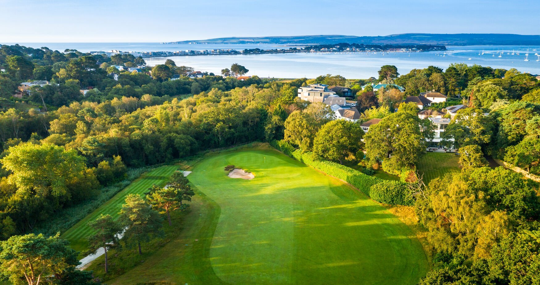 Aerial view of a lush green golf course surrounded by trees with bay and houses visible in the background