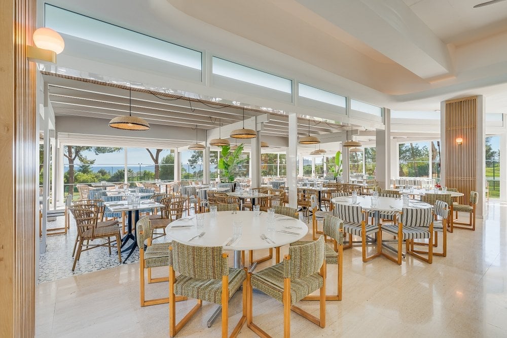 Modern restaurant dining room with round white tables, wooden chairs, and floor-to-ceiling windows overlooking outdoor seating area