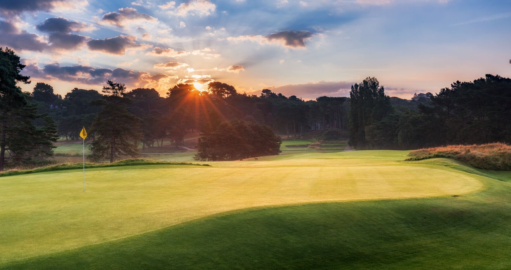 Golf course fairway at sunset with manicured green grass, trees lining the course, and golden sky with clouds