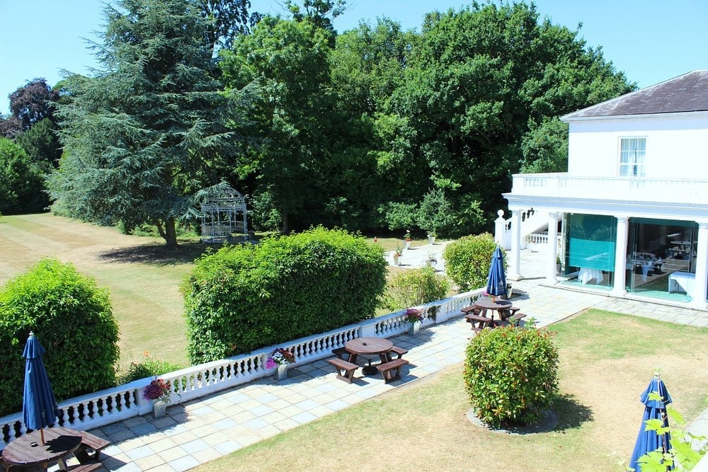 Aerial view of a white house with terrace, manicured hedges, and patio area surrounded by trees on a sunny day
