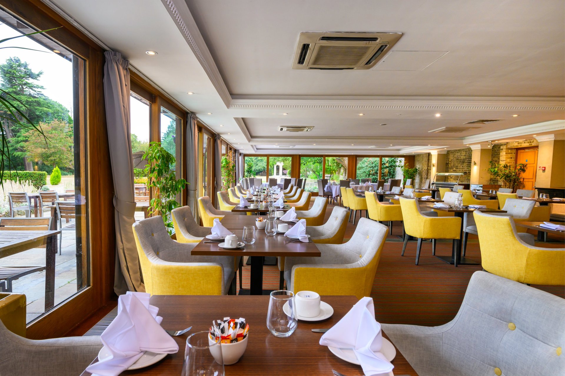 Upscale restaurant dining room with yellow chairs, black tables, and floor-to-ceiling windows overlooking a garden patio