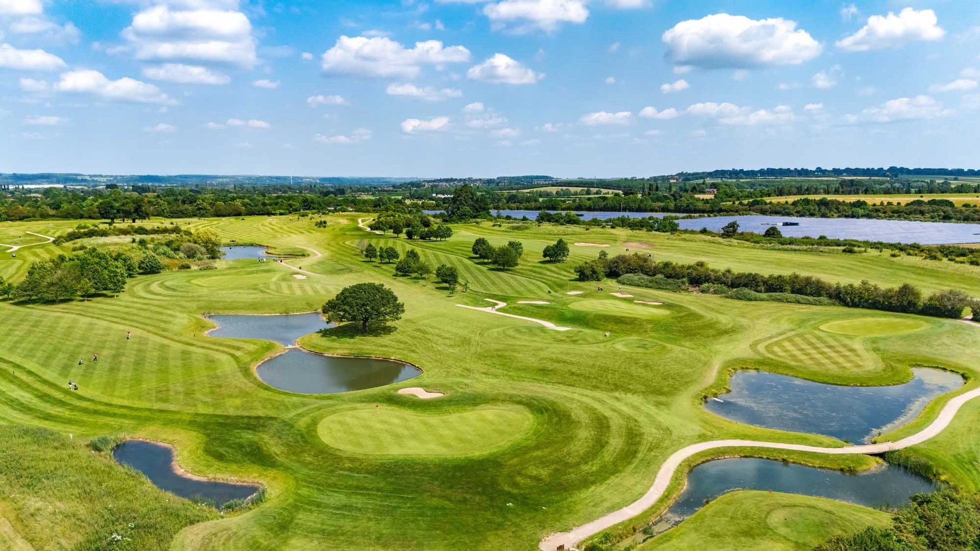 Aerial view of a scenic golf course with manicured fairways, water hazards, and surrounding countryside under blue sky