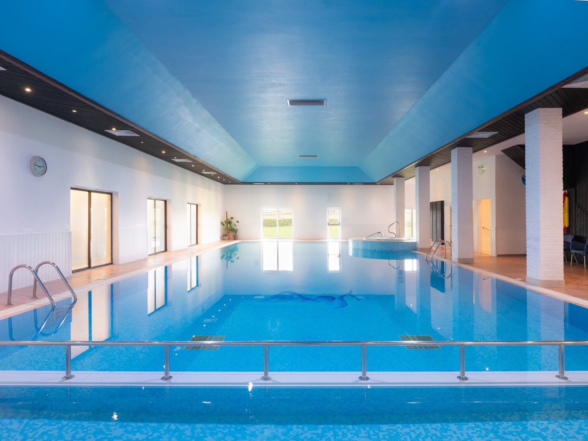 Indoor swimming pool with blue water, metal safety railings, white columns, and bright blue ceiling in modern facility