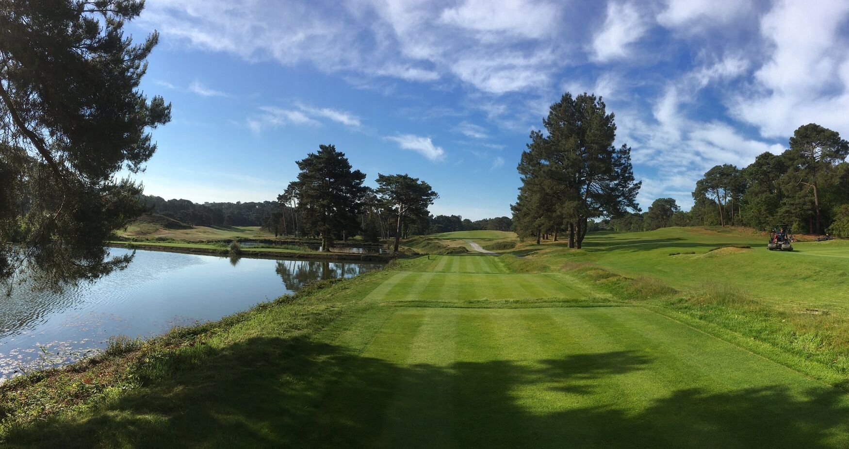Golf course fairway with manicured green grass, water hazard on left, trees lining the course under blue sky with clouds