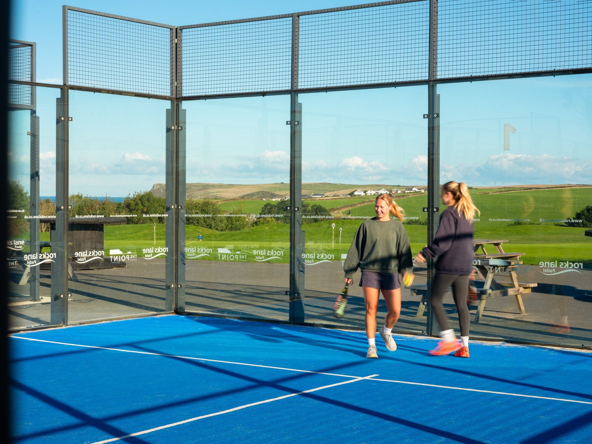Two people standing on a blue outdoor sports court with protective netting, overlooking green fields and sky