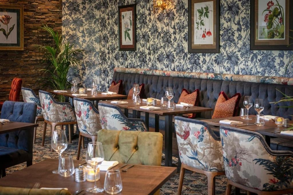 Upscale restaurant dining room with blue floral wallpaper, botanical framed art, booth seating with patterned cushions, wood tables, and water glasses