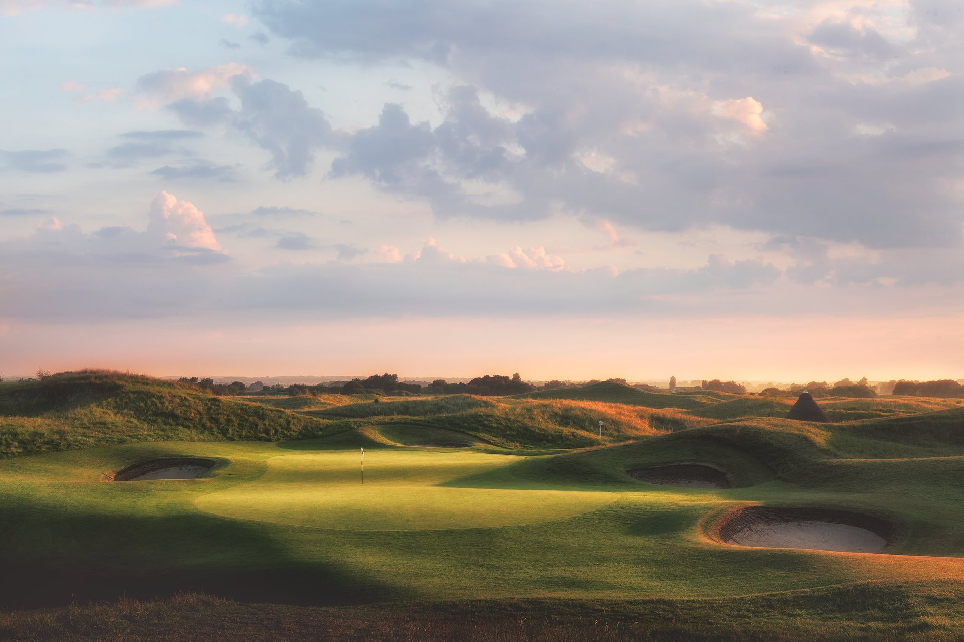 Rolling hills golf course landscape with bunkers and water hazard at sunset under cloudy sky