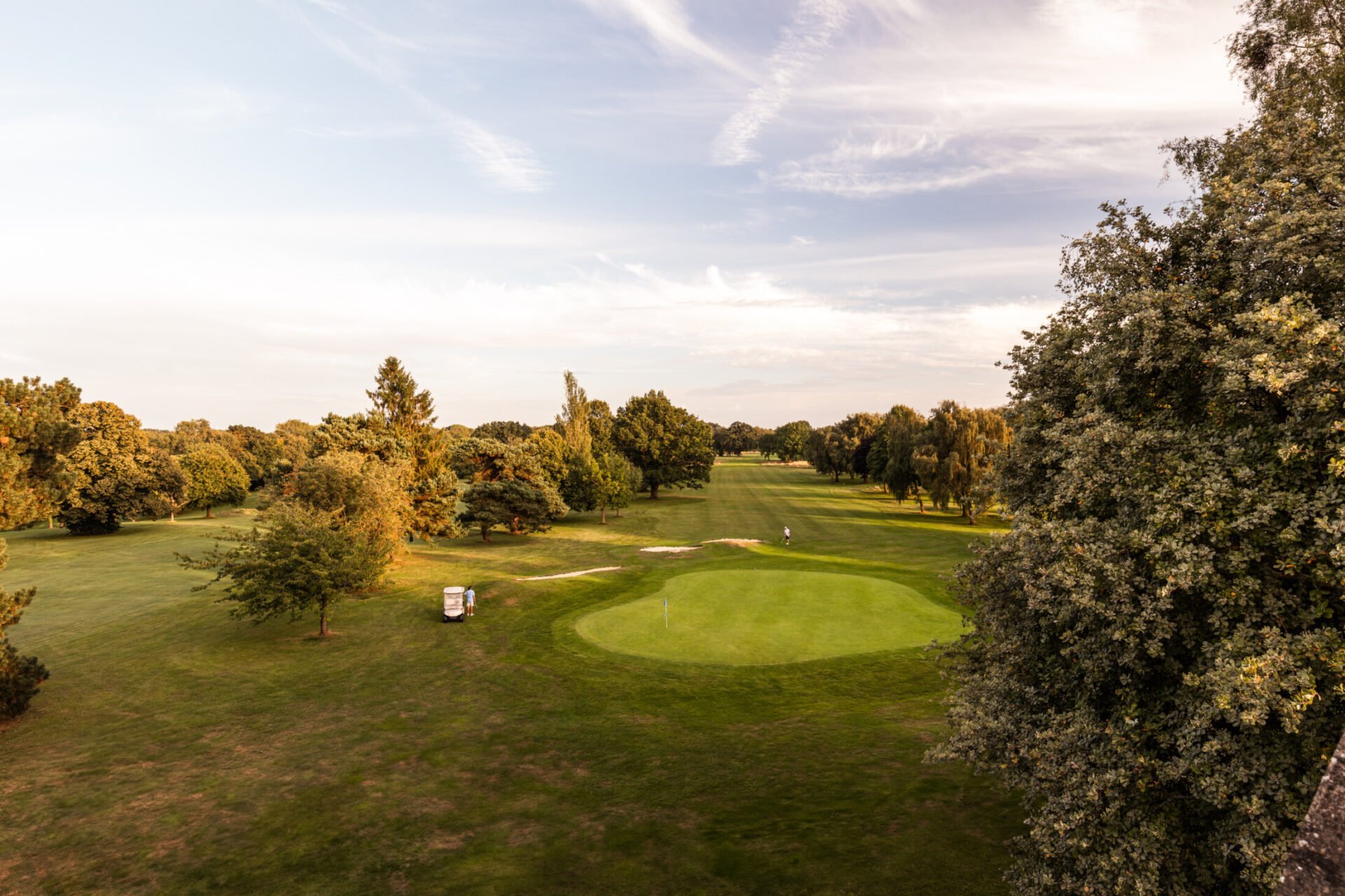 Aerial view of a golf course green with fairway, surrounded by mature trees and a clubhouse, under a cloudy sky at sunset