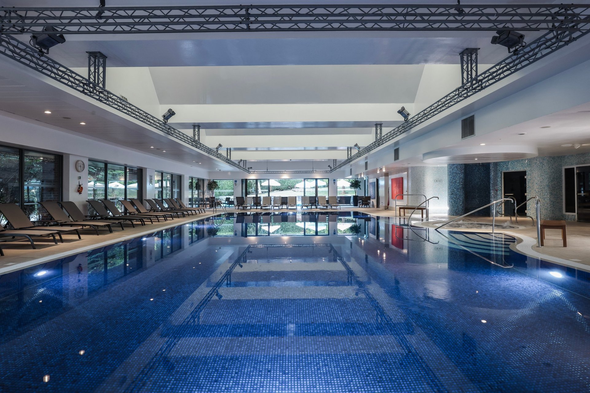 Indoor Olympic-sized swimming pool with lane dividers, high ceiling with industrial trusses, lounge seating, and modern facility lighting
