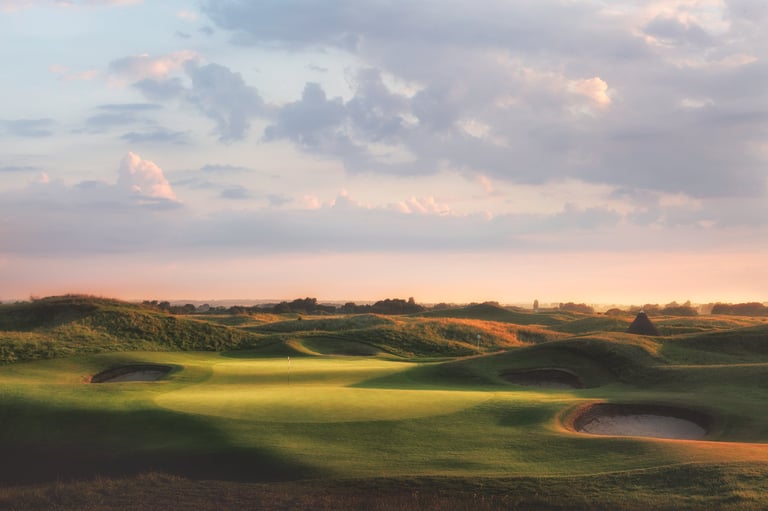 Rolling green golf course with sand bunkers and water hazard at sunset under dramatic cloudy sky