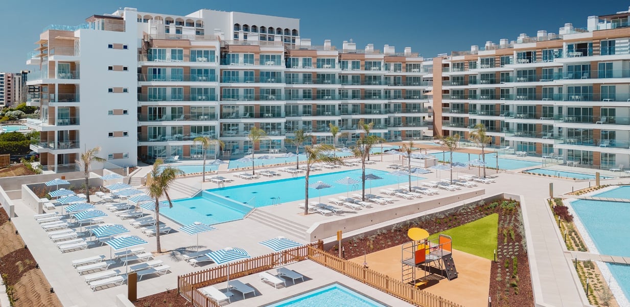 Luxury beachfront resort with modern apartment buildings overlooking multiple swimming pools and sandy beach under clear blue sky