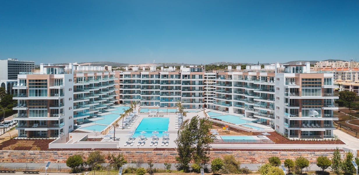 Aerial view of a modern residential complex with curved white buildings surrounding a blue swimming pool and recreational area