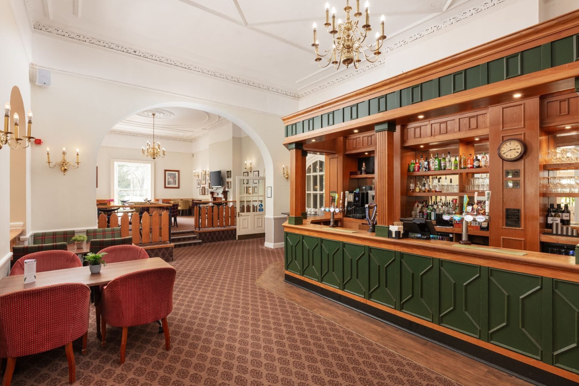 Elegant hotel lobby with burgundy carpeting, red armchairs, wooden bar counter with green paneling, brass chandelier, and arched ceiling